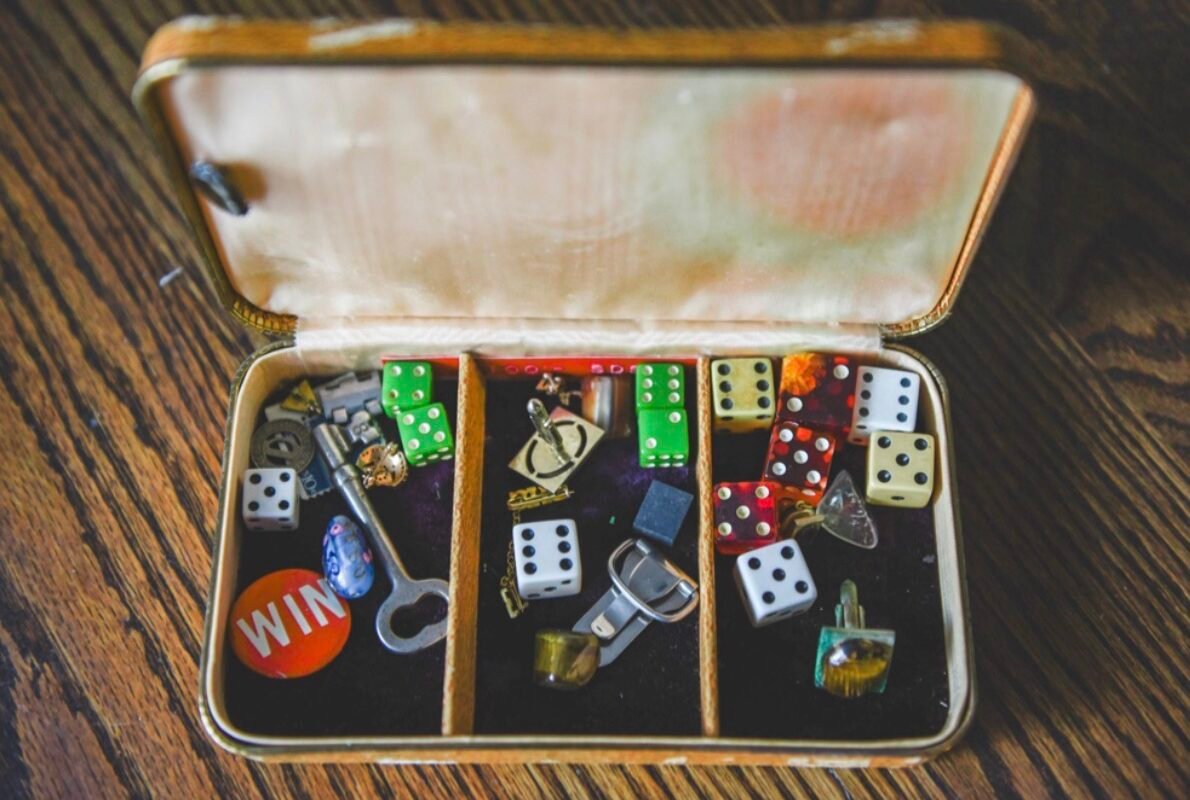 An open vintage box sitting on a wooden surface, filled with colorful gaming dice, cufflinks, a key, and a pin.